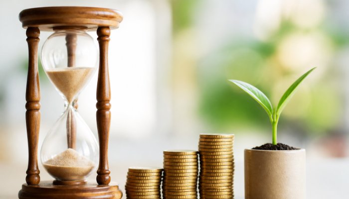 A close-up of a wooden hourglass next to a stack of gold coins and a green sprout growing out of a small pot, signifying