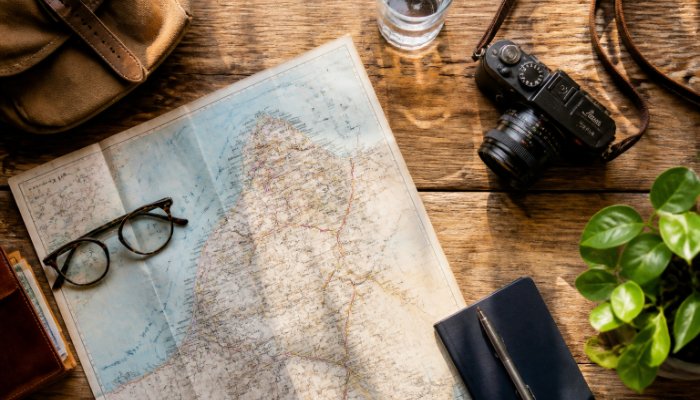 An overhead shot of an organized travel flat-lay including a map, a high-end camera, glasses, and a glass of water on a